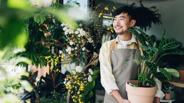 confident-young-asian-male-florist-owner-of-small-business-flower-shop-holding-potted-plant-outside-his-workplace-he-is-looking-away-with-smile
